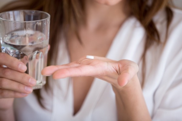 Auf einer ausgestreckten Damenhand liegt eine Tablette. In der anderen Hand wird ein Glas mit Wasser gehalten.