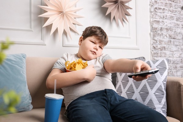Kind mit Übergewicht sitzt mit Snacks vor dem Fernseher