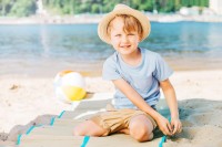 Kleiner Junge sitzt mit Hut bedeckt im Schatten an einem See.